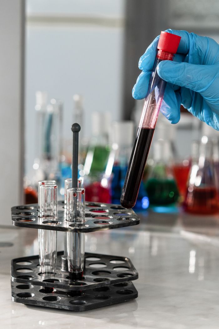 Gloved hand holding test tube in a laboratory setting with colorful beakers in background.